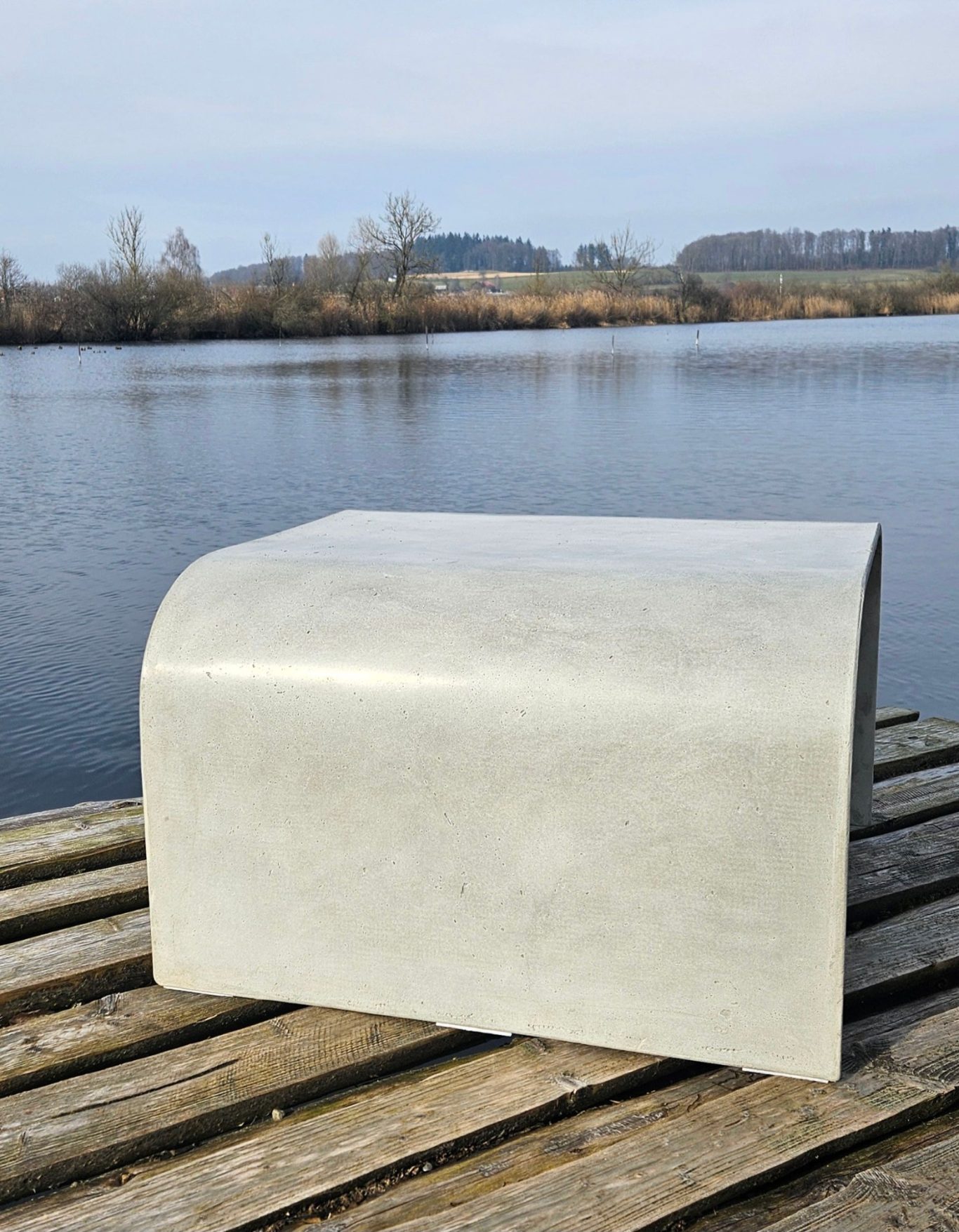 Tavolo Bene 1 Moderner, grauer Hocker aus Beton auf einem Holzsteg an einem ruhigen Gewässer.